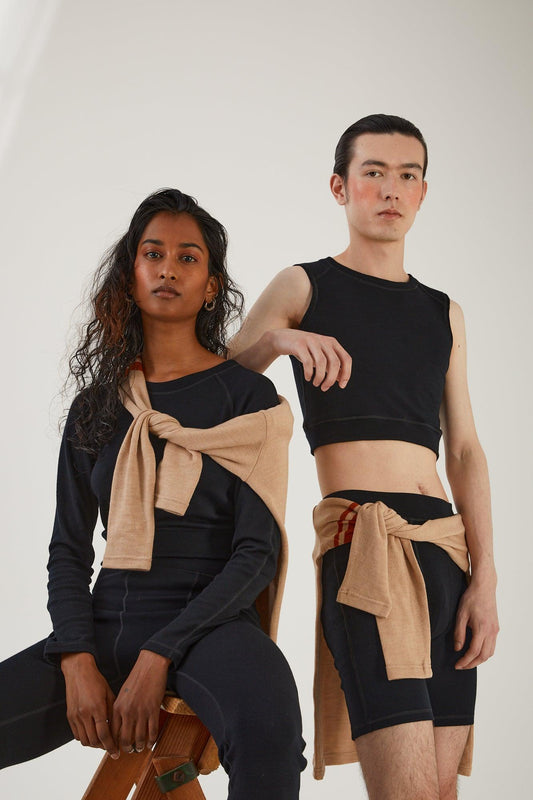 A person sitting on a wooden step ladder wearing merino wool leggings and a merino wool cropped long sleeve top with a merino wool jumper draped around their shoulders next to a person standing wearing merino wool biker shorts and merino wool cropped tank with a merino wool jumper tied around their waist