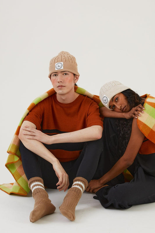Two people sitting on the ground resting against each other, both wearing merino wool beanies, a merino wool top, merino wool leggings, merino wool socks, merino wool track pants and a merino wool vest with a wool blanket draped over their shoulders